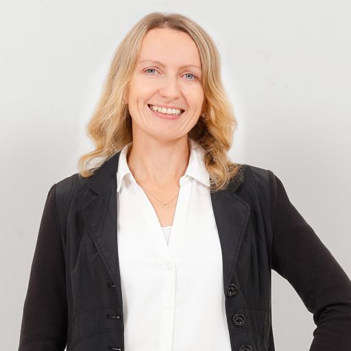 Lächelnde Frau mit blonden Haaren im schwarzen Blazer und weißer Bluse vor hellem Hintergrund.
