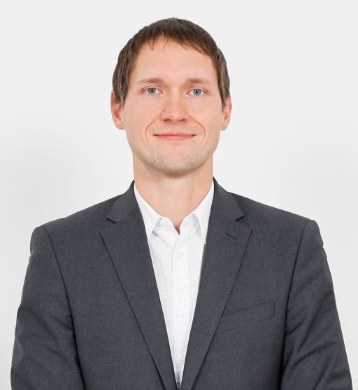 Mann mit kurzen braunen Haaren und Dreitagebart im grauen Sakko über weißem Hemd vor weißem Hintergrund.