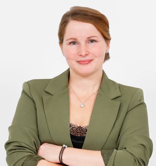 Frau mit roten Haaren, grünem Blazer und verschränkten Armen vor weißem Hintergrund, lächelt in die Kamera.