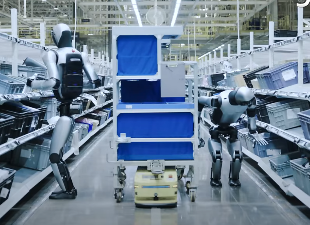 Zwei humanoide Roboter stehen in einem Lagergang zwischen Regalen, dahinter ein fahrerloses Transportfahrzeug mit blauem Regal.