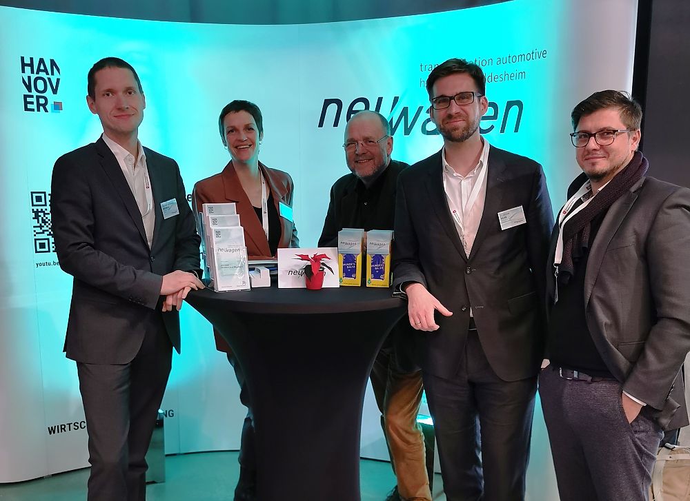 Fünf Personen stehen um einen Stehtisch vor einem Messebanner mit den Aufschriften „Hannover" und „newwagen".