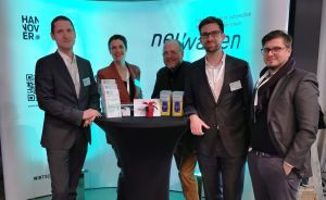 Fünf Personen stehen um einen Stehtisch vor einem Messebanner mit den Aufschriften „Hannover" und „newwagen".